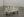 A minimalist sculpture by Brittany Mojo featuring a small cart with wheels, painted entirely in white, carrying an assortment of white-painted bottles and containers. Displayed in La Sierra Art gallery with gray flooring and plain walls, the piece has a soft, ghostly look.