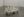 A minimalist sculpture by Brittany Mojo featuring a small cart with wheels, painted entirely in white, carrying an assortment of white-painted bottles and containers. Displayed in La Sierra Art gallery with gray flooring and plain walls, the piece has a soft, ghostly look.