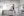 An installation space at La Sierra University Brandstater Gallery by Brittany Mojo, with minimalist drawings covering all walls. The room's center has wooden sawhorses and scattered items, adding a workshop vibe, while a shelf above holds bottles and a plant, contrasting with the clean, ordered art display